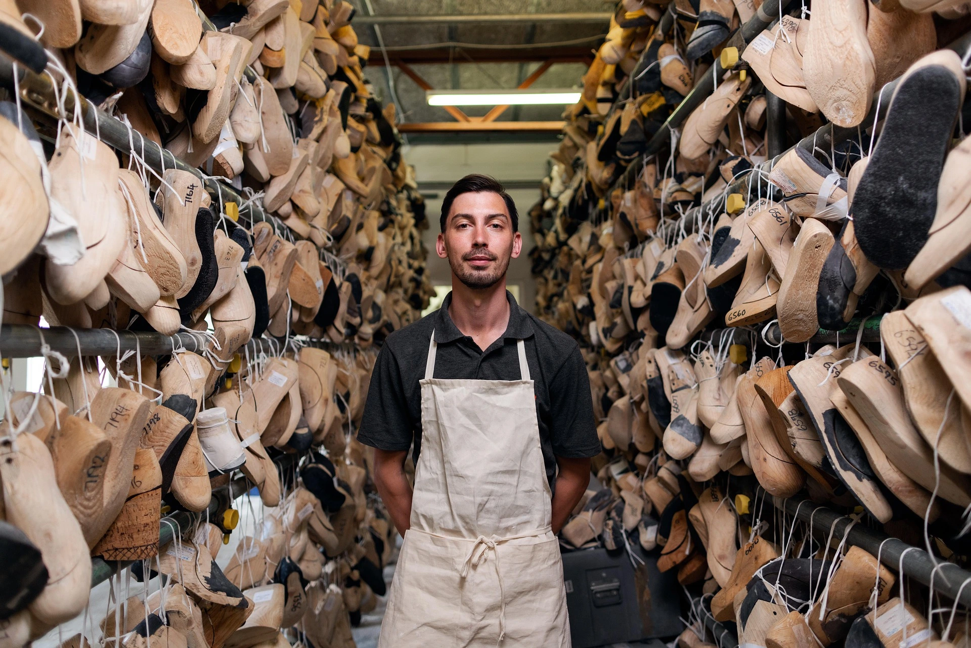 Man wearing a beige apron standing in a workshop aisle filled with wooden shoe molds