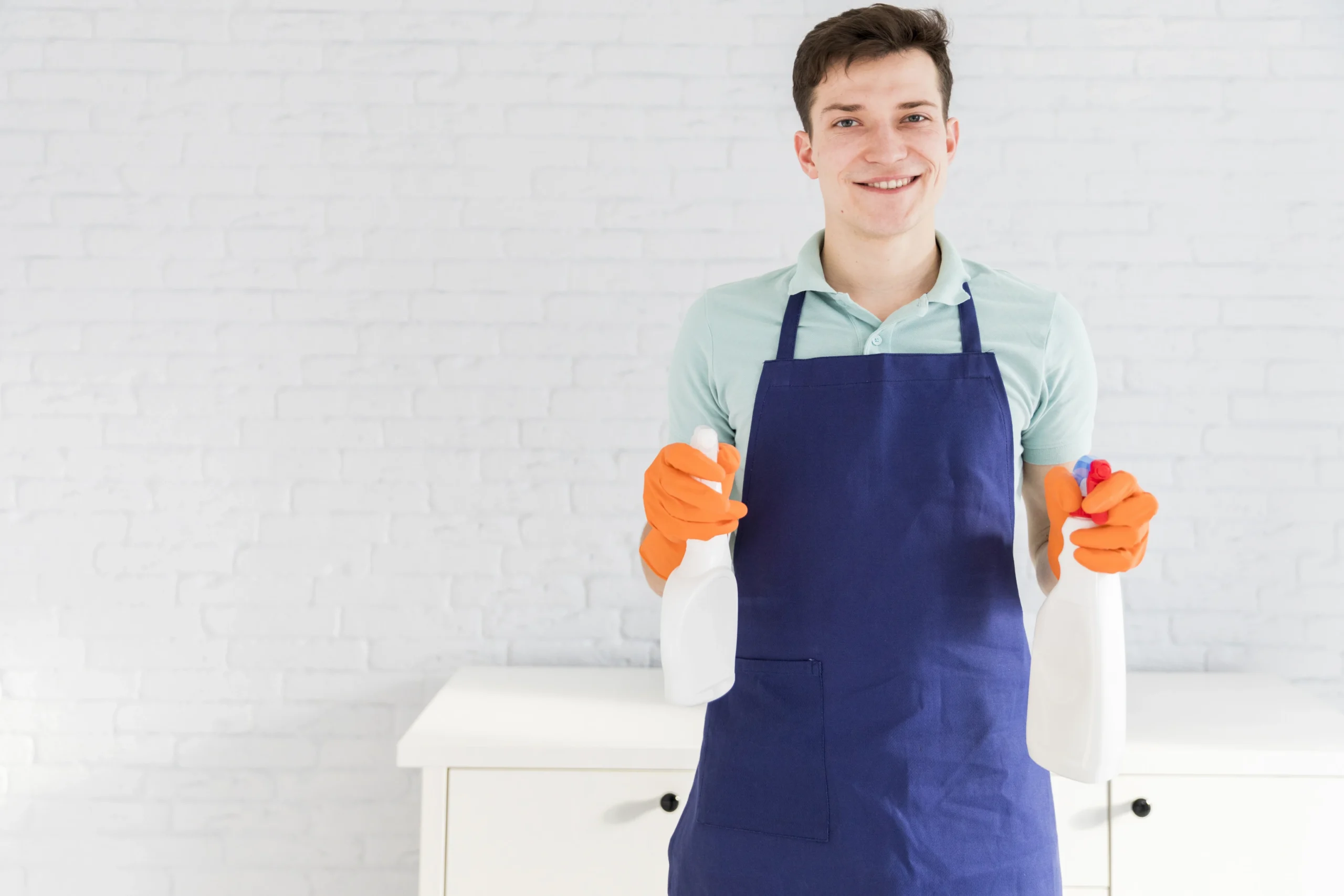 Smiling man in a blue apron holding cleaning spray bottles and wearing orange gloves